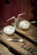 The Vagabond Vintage Stork Salt and Pepper Cellar features glass inserts and silver spoons joined by a stork figurine, displayed on a rustic wooden crate marked 1917.