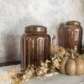 A Ragon House 11" Amber Glass Jar with an antiqued lid features dried flowers and wooden pumpkins, displayed on a light cloth before a textured gray wall; another matching jar is softly blurred in the background.