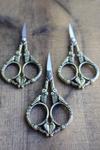 Three pairs of NNK Press Feathered Friends Scissors: Antique Gold, featuring intricate handles and stainless steel blades, are arranged on a wooden surface with blades pointing outward.