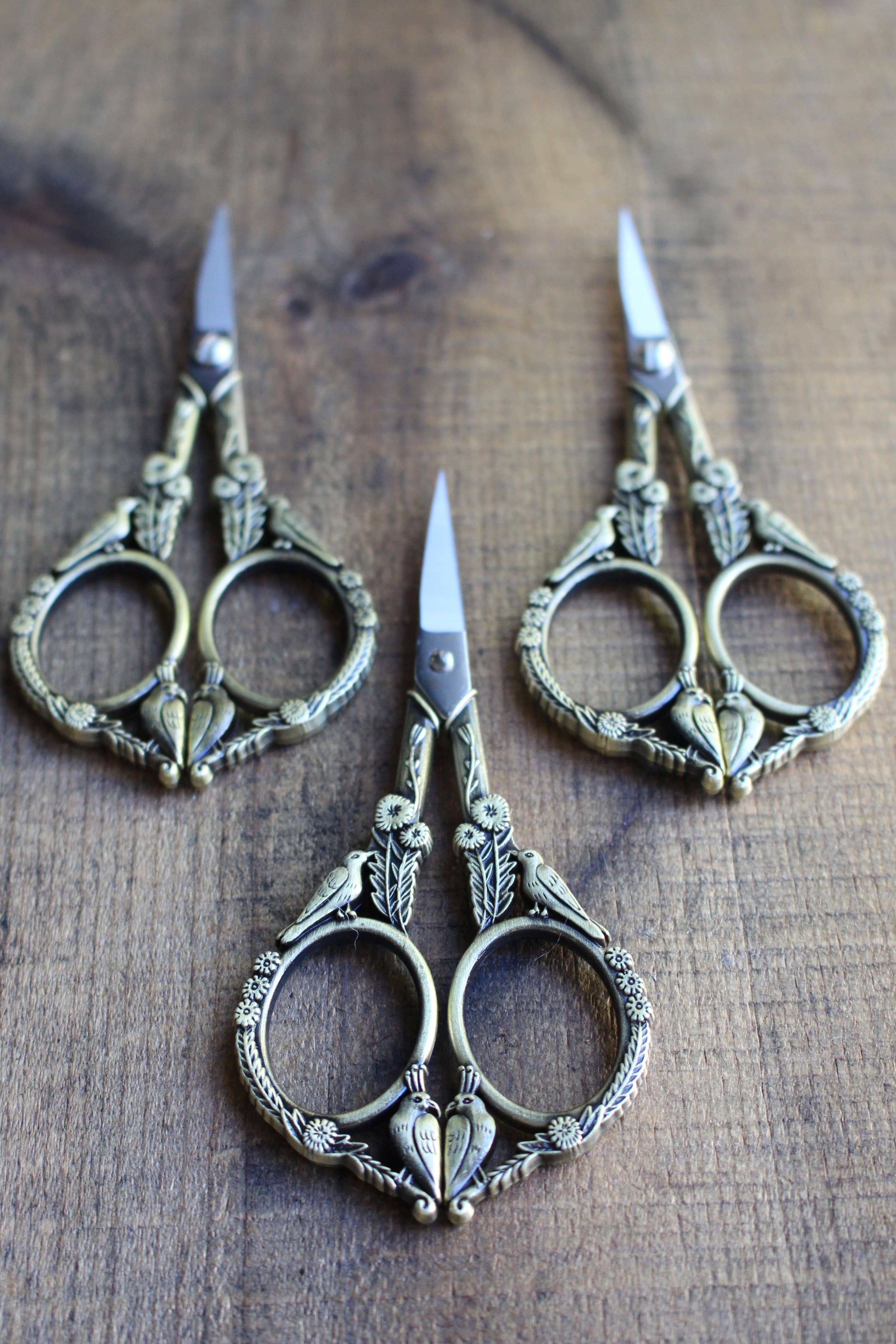 Three pairs of NNK Press Feathered Friends Scissors: Antique Gold, featuring intricate handles and stainless steel blades, are arranged on a wooden surface with blades pointing outward.