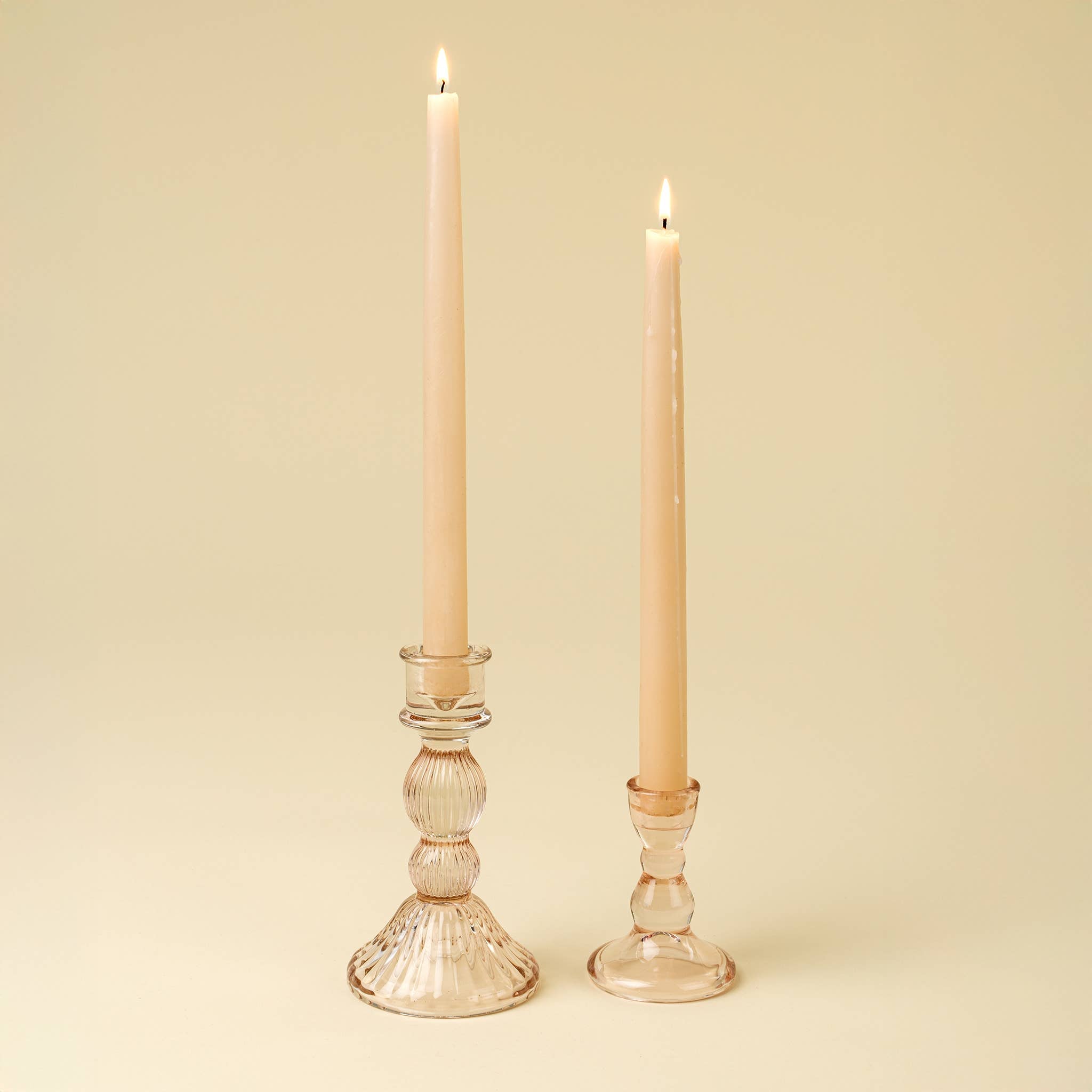 Two lit cream-colored taper candles sit in clear glass holders against a light beige background. The taller, more ornate holder is the Callista Glass Candleholder Large in Blush by Indaba Trading, adding a vintage-inspired touch to the display.