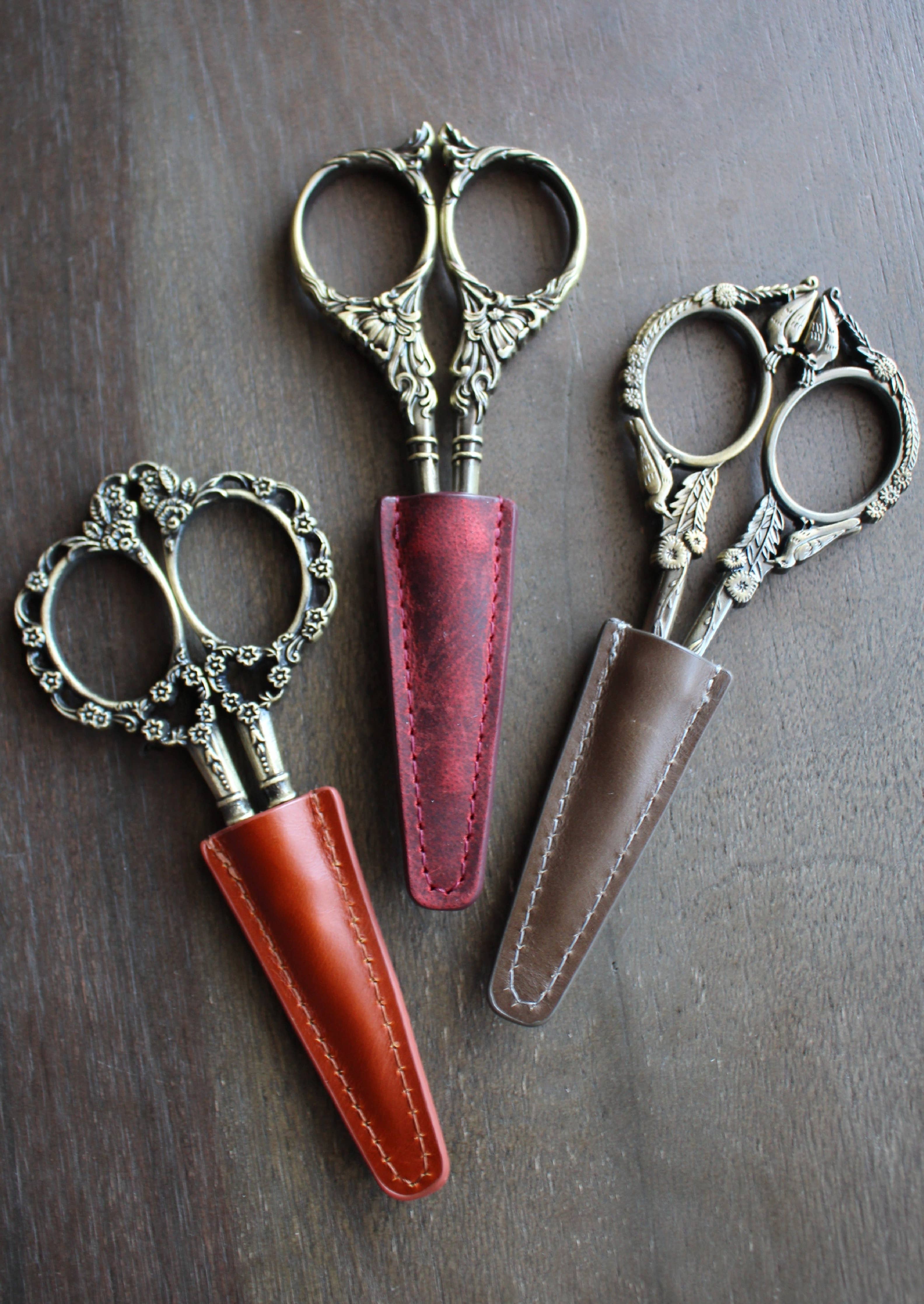 Three embroidery scissors with intricate handles sit in red, brown, and orange NNK Press Leather Scissor Sheaths, fanned out on a wooden surface—perfect as unique embroidery accessories.
