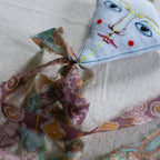 The Flying Kite - Cotton & Lavender filled Ornament by Skippy Cotton is handmade from organic cotton with a painted face—blue eyes, red cheeks, stitched yellow lines—and features a patterned ribbon tail, resting on a cream textured surface.