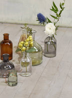 A collection of Jamali Garden vases, including the Smoke Medicine Bottle Bud Vase, displayed on a light wooden floor. The glass vases vary in size, shape, and color—clear, smoke-grey, and amber—and hold flowers or greenery.