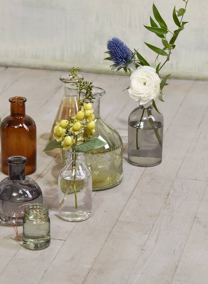 A collection of Jamali Garden vases, including the Smoke Medicine Bottle Bud Vase, displayed on a light wooden floor. The glass vases vary in size, shape, and color—clear, smoke-grey, and amber—and hold flowers or greenery.