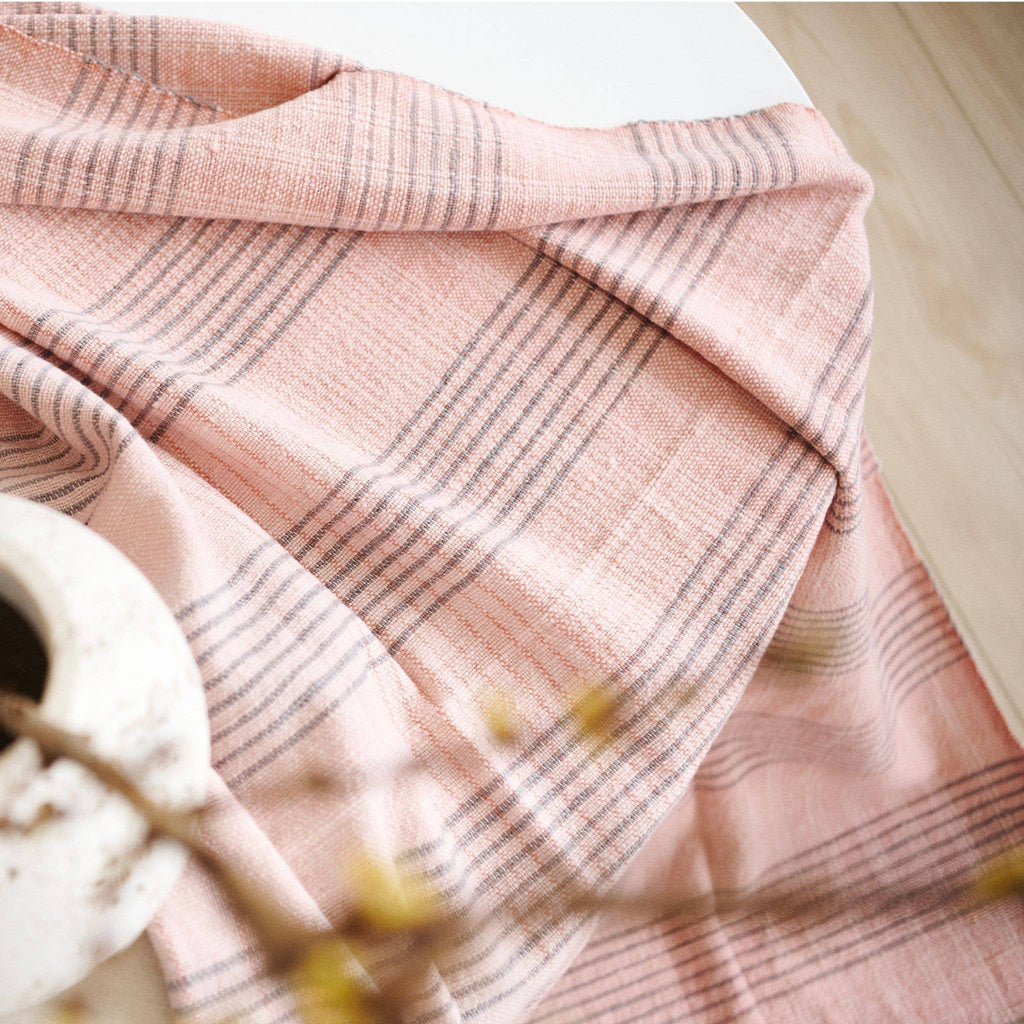 A Silica Rose Throw in shades of salmon and gray stripes by Variously is draped across a light surface, with part of a white ceramic vase and blurred yellow flowers in the foreground.