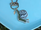 The Metal Enamel Keychain / Purse Charm | Snail by ilikesara | art + goods features a charming metal snail and leaf-shaped charm, silver plated, resting on a light blue surface alongside vintage keys.
