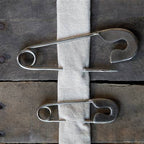 Two HomArt Safety Pin - Lrg - Nickel secure a strip of white fabric to a rustic, weathered wooden surface, creating a unique and stylish key holder.