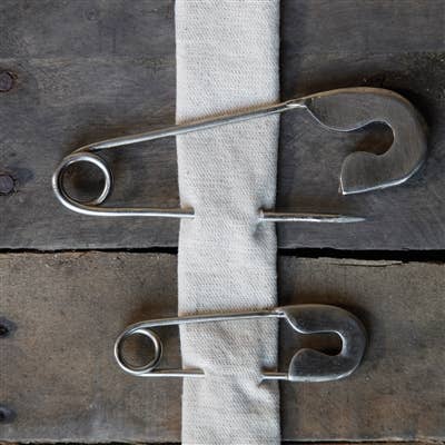 Two HomArt Safety Pin - Lrg - Nickel secure a strip of white fabric to a rustic, weathered wooden surface, creating a unique and stylish key holder.