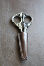 The NNK Press Leather Scissor Sheath protects your embroidery scissors with style—an elegant accessory that keeps your ornate tools safe and organized on any crafting surface.