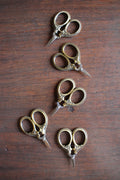 Four pairs of NNK Press Mini Embroidery Scissors in Antique Gold, featuring stainless steel blades and gold handles, are displayed in a staggered vertical line on a wooden surface.