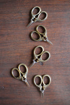 Four pairs of NNK Press Mini Embroidery Scissors in Antique Gold, featuring stainless steel blades and gold handles, are displayed in a staggered vertical line on a wooden surface.
