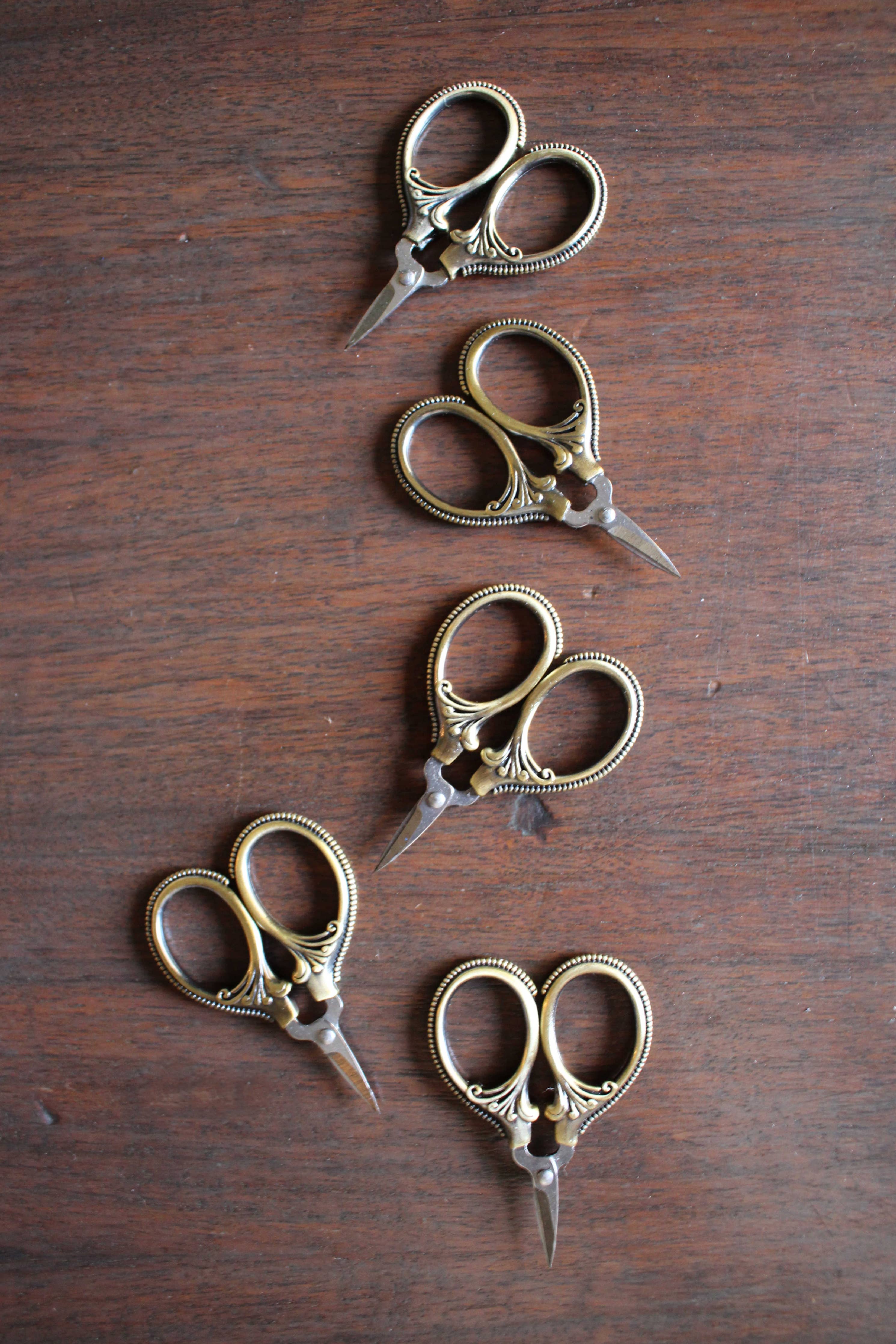 Four pairs of NNK Press Mini Embroidery Scissors in Antique Gold, featuring stainless steel blades and gold handles, are displayed in a staggered vertical line on a wooden surface.