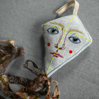 The Skippy Cotton Flying Kite ornament, crafted from organic cotton and lavender-filled for a scented touch, features an embroidered face and floral tail—perfect as a charming gift topper on any gray textured surface.
