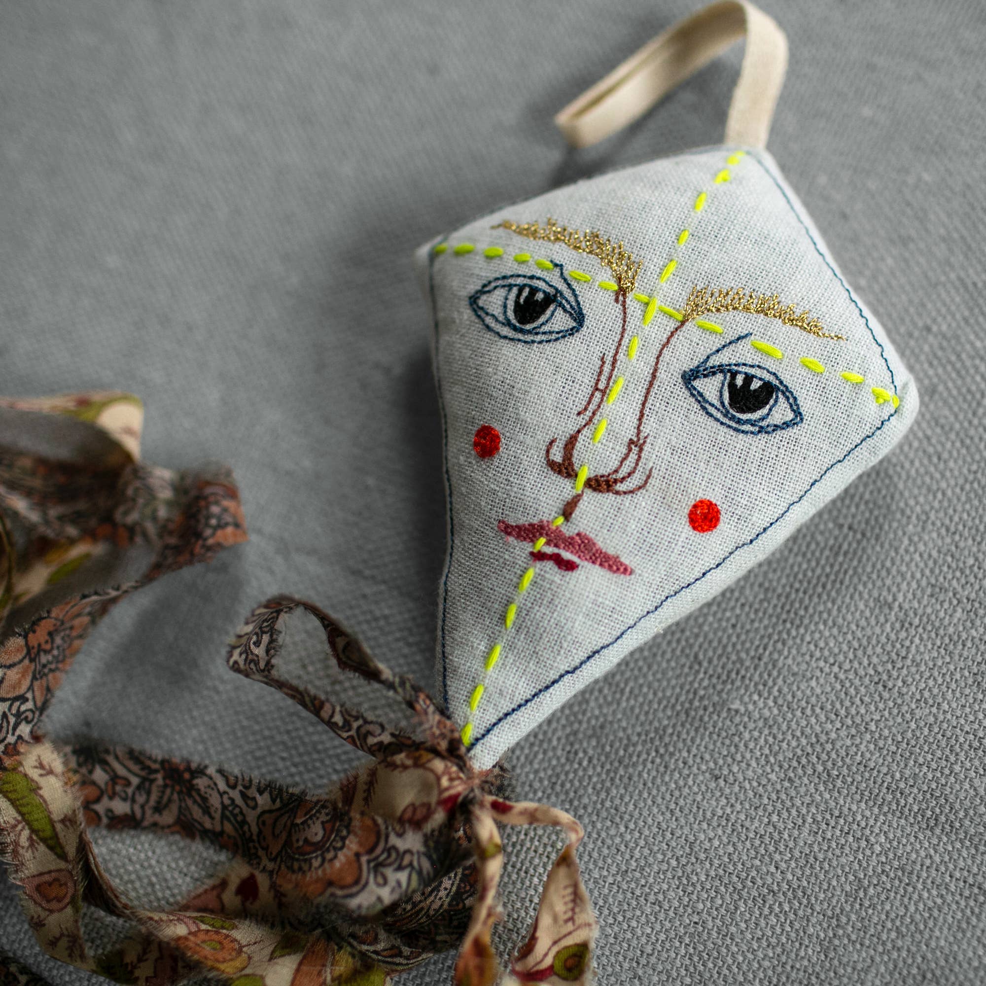 The Skippy Cotton Flying Kite ornament, crafted from organic cotton and lavender-filled for a scented touch, features an embroidered face and floral tail—perfect as a charming gift topper on any gray textured surface.