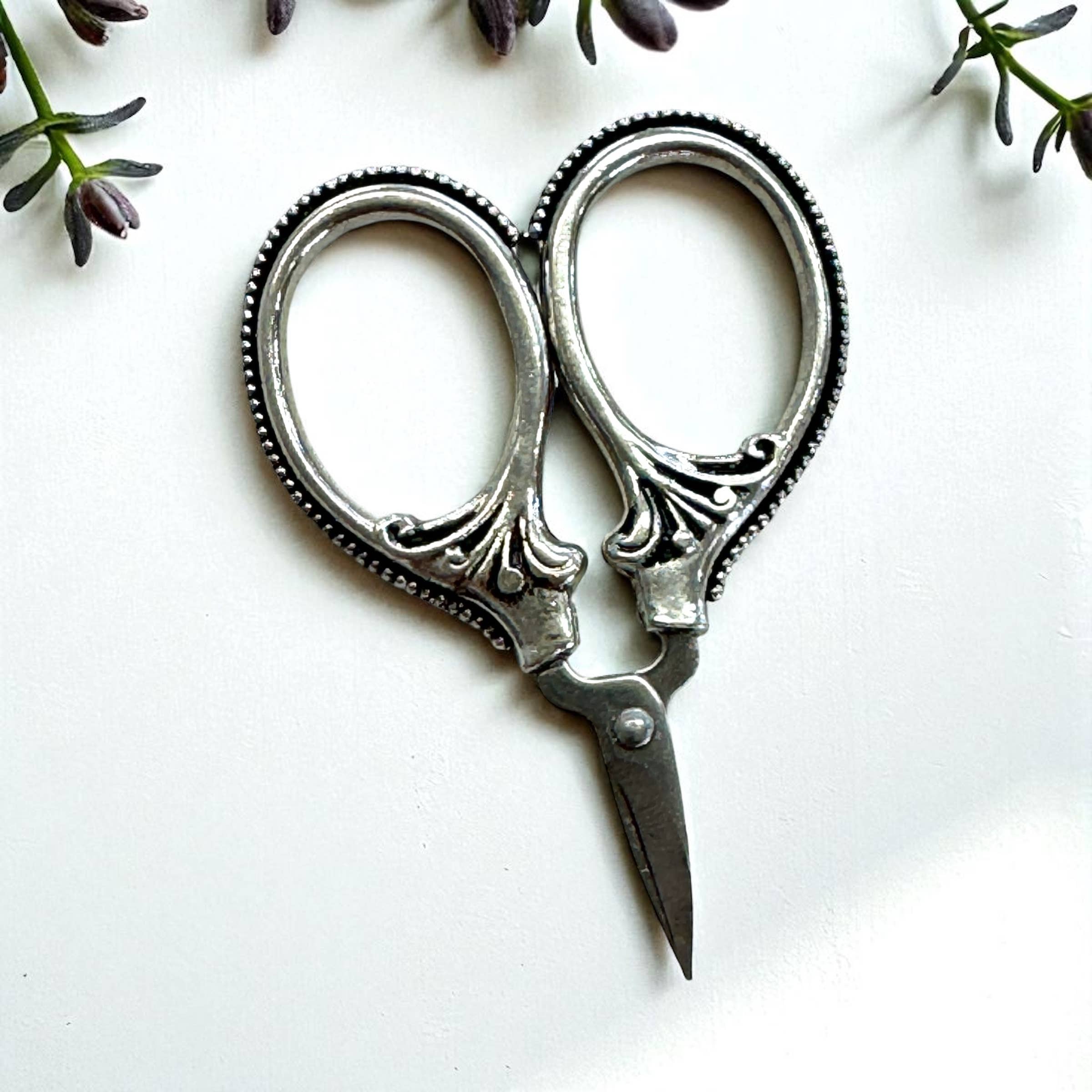 Firefly Notes' Petite Thread Snips, vintage-style portable sewing scissors with decorative handles, rest on a white surface bordered by sprigs of small purple flowers.