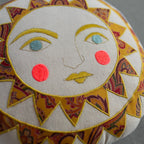 The Skippy Cotton Bright Neon Sun Pillow is a handmade, cotton-filled cushion with an embroidered sun face—yellow rays, teal eyes, yellow lips, and pink cheeks on beige organic cotton—perfect for whimsical decor in any space.
