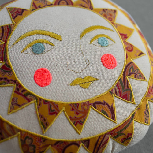 The Skippy Cotton Bright Neon Sun Pillow is a handmade, cotton-filled cushion with an embroidered sun face—yellow rays, teal eyes, yellow lips, and pink cheeks on beige organic cotton—perfect for whimsical decor in any space.