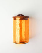 The Handblown Hammered Glass Canisters: Smoke / Small by Creative Women is a tall smoke glass canister with a round hand-carved wooden lid and handle, featuring an airtight seal, displayed on a white surface.