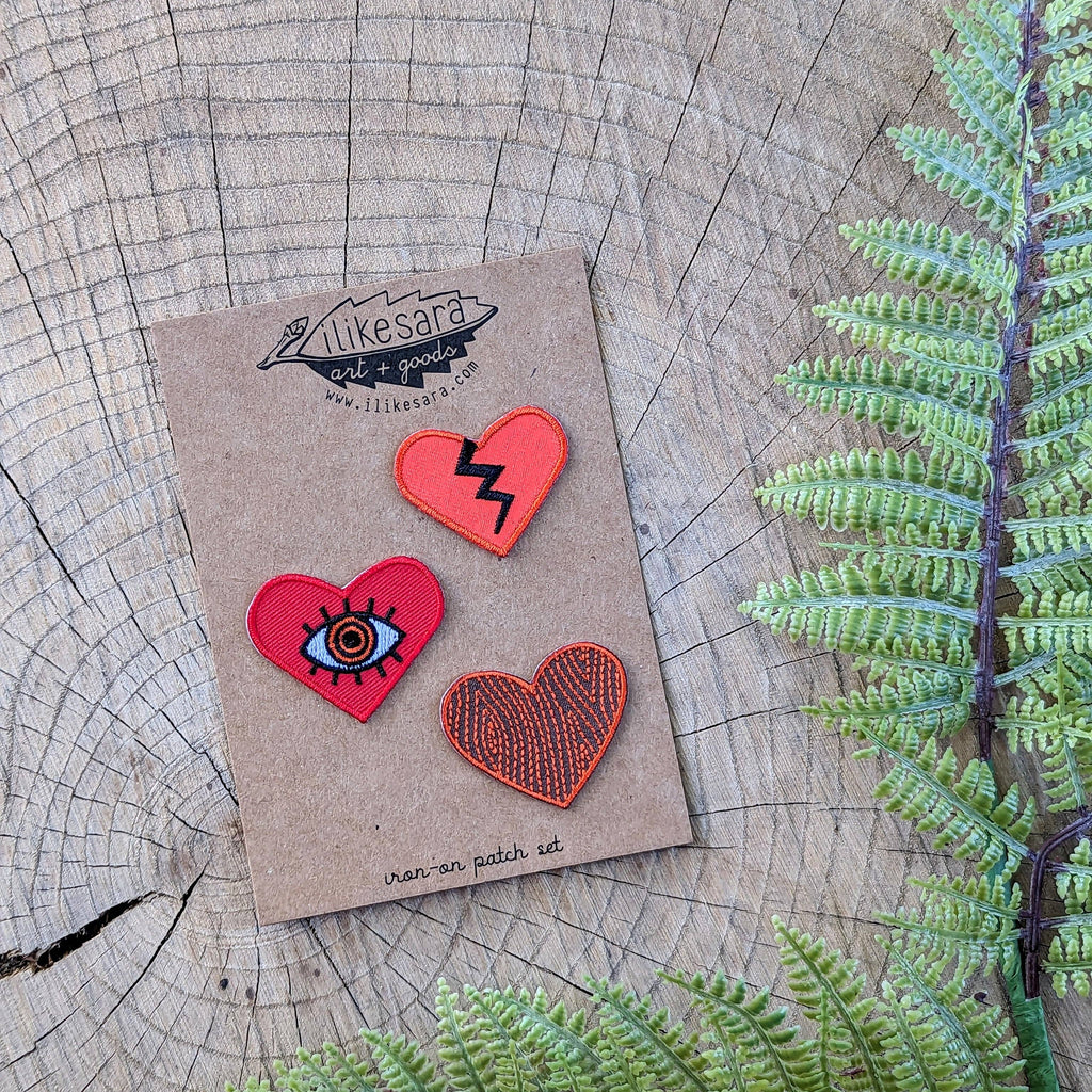 The ilikesara | art + goods Iron-on patch | heart 3-pack - red features three unique embroidered heart patches, each with a distinct design, displayed on a brown card alongside green fern leaves.