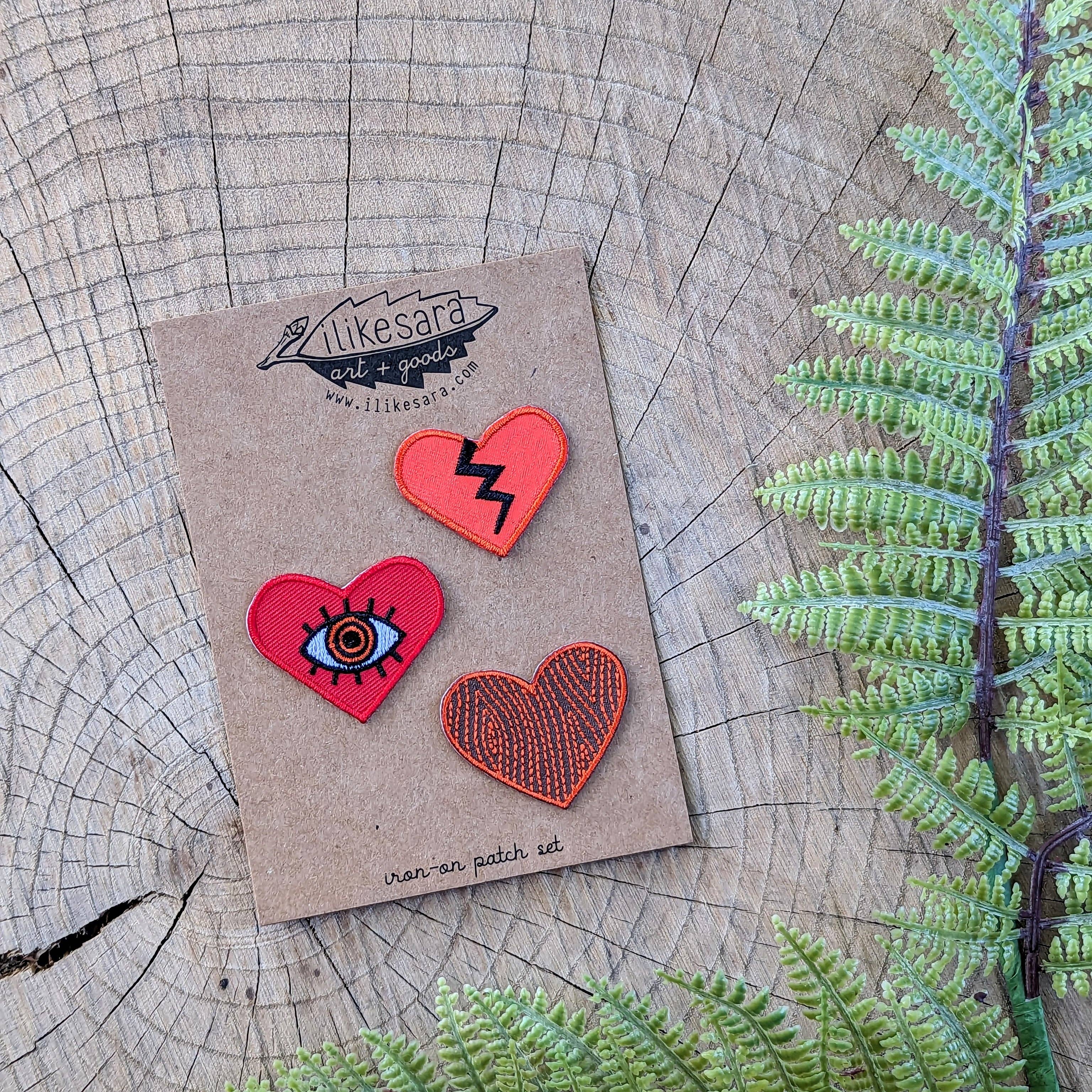 The ilikesara | art + goods Iron-on patch | heart 3-pack - red features three unique embroidered heart patches, each with a distinct design, displayed on a brown card alongside green fern leaves.