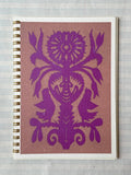 The Parcel Papercut Flower Notebook is a 5x7 spiral-bound notebook with sturdy cardstock cover, adorned with a symmetrical purple folk-art bird and flower design on brown, shown resting on lined paper.