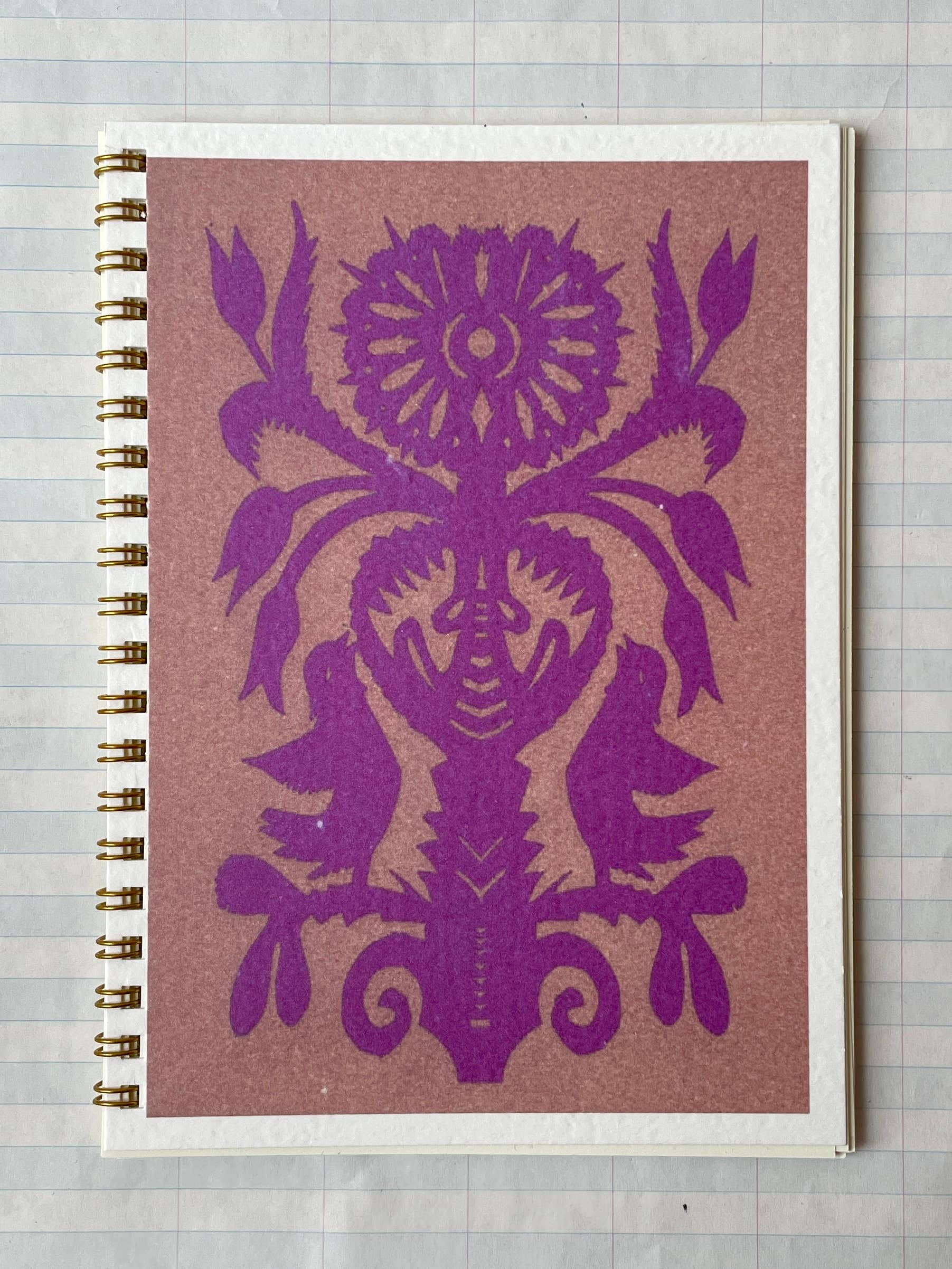 The Parcel Papercut Flower Notebook is a 5x7 spiral-bound notebook with sturdy cardstock cover, adorned with a symmetrical purple folk-art bird and flower design on brown, shown resting on lined paper.