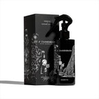 A black O TANNENBAUM ROOM SPRAY: 200ML by Heretic Parfum stands beside its matching box, both adorned with whimsical white figures and a large tree—evoking Edward Gorey's style and perfect for a home fragrance with fir needle notes.