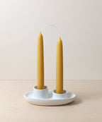 A pair of 7-inch hand-dipped pure beeswax tapers by Yarrow Bone Farmstead, joined by an uncut wick, stand upright in a white ceramic holder with two slots on a light wooden surface against a neutral background.