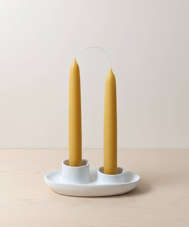 A pair of 7-inch hand-dipped pure beeswax tapers by Yarrow Bone Farmstead, joined by an uncut wick, stand upright in a white ceramic holder with two slots on a light wooden surface against a neutral background.