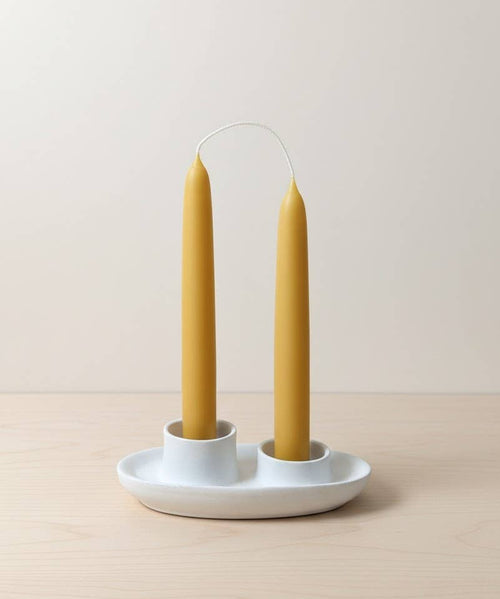 A pair of 7-inch hand-dipped pure beeswax tapers by Yarrow Bone Farmstead, joined by an uncut wick, stand upright in a white ceramic holder with two slots on a light wooden surface against a neutral background.