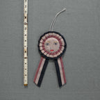 The Skippy Cotton Prize Ribbon is a handmade cotton-filled ornament with an embroidered face, red lips, and ribbon tails, measuring about 18 cm long. It’s shown next to a measuring tape on a grey textured surface.