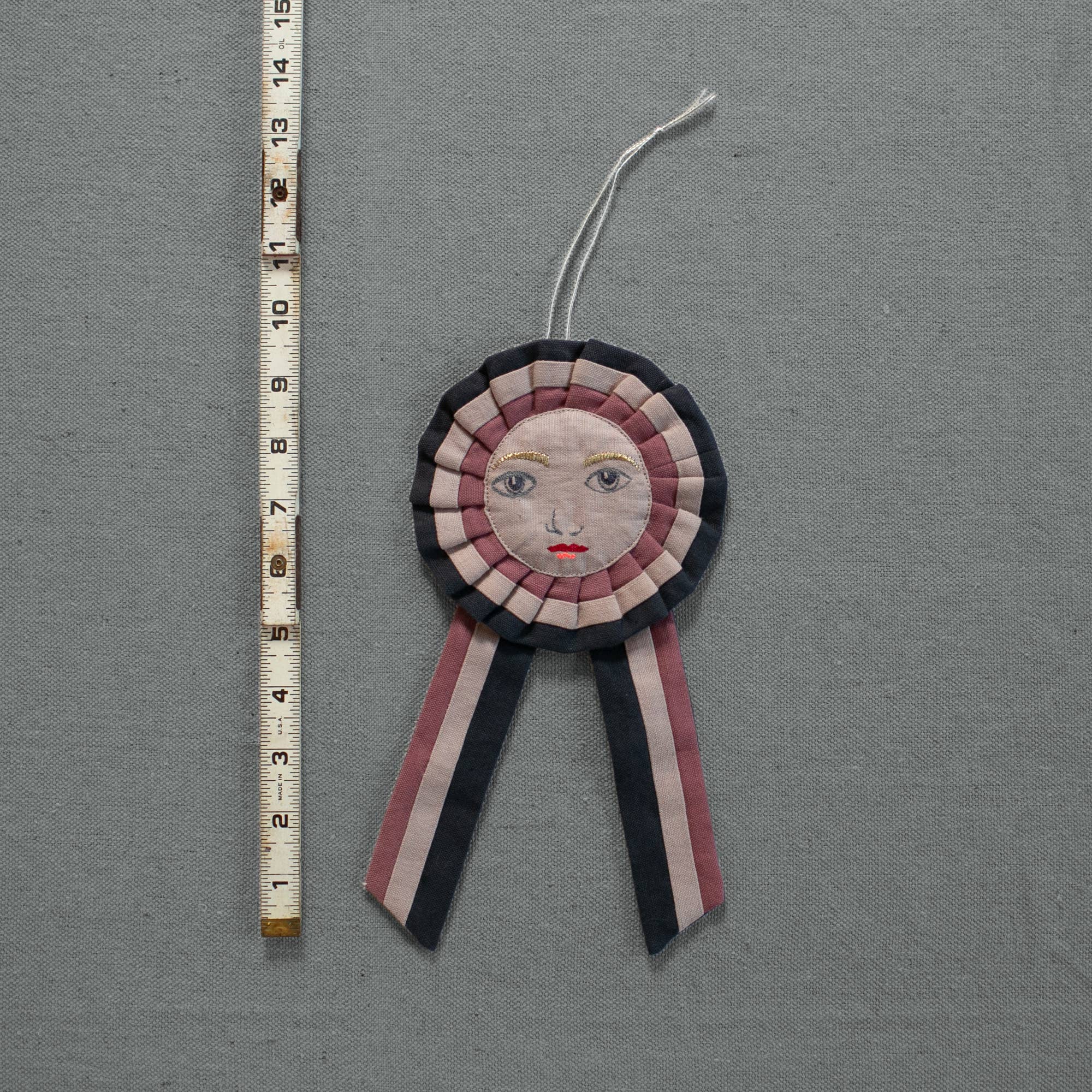 The Skippy Cotton Prize Ribbon is a handmade cotton-filled ornament with an embroidered face, red lips, and ribbon tails, measuring about 18 cm long. It’s shown next to a measuring tape on a grey textured surface.