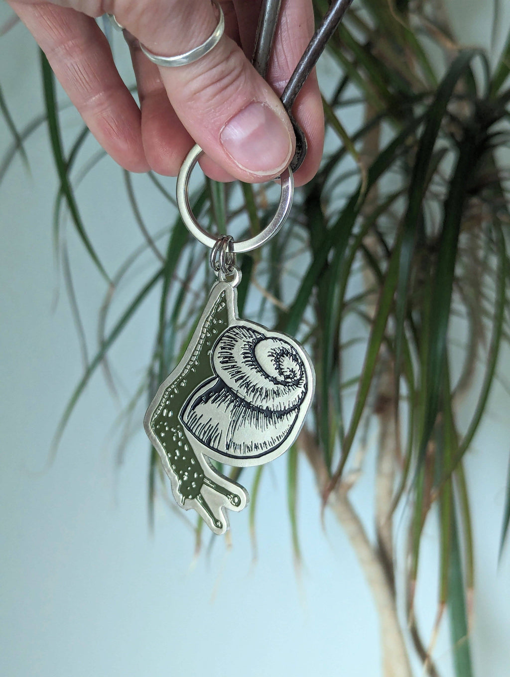 A hand holds the ilikesara | art + goods Metal enamel keychain / purse charm featuring a cute snail design, with a leafy houseplant softly blurred in the background.