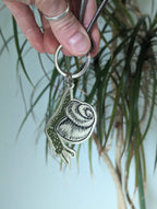 A hand holds the ilikesara | art + goods Metal enamel keychain / purse charm featuring a cute snail design, with a leafy houseplant softly blurred in the background.