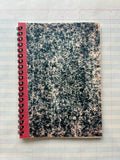 The Parcel Antique Endpaper Notebook: Small - Composition / Unlined, featuring a marbled black-and-white cover with a red spine, rests on blue and pink grid graph paper next to a 5x7 printed card.