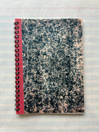 The Parcel Antique Endpaper Notebook: Small - Composition / Unlined, featuring a marbled black-and-white cover with a red spine, rests on blue and pink grid graph paper next to a 5x7 printed card.