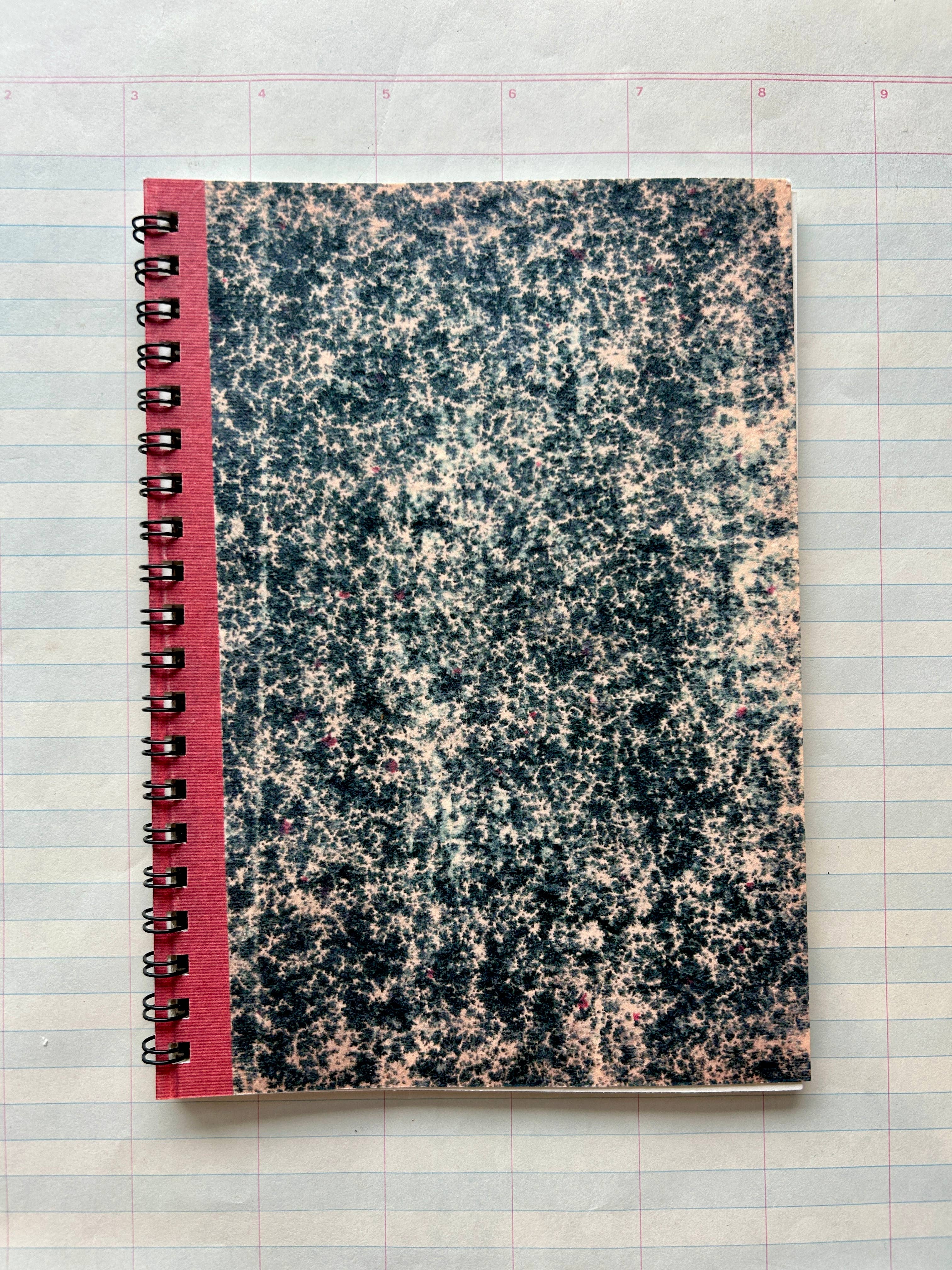 The Parcel Antique Endpaper Notebook: Small - Composition / Unlined, featuring a marbled black-and-white cover with a red spine, rests on blue and pink grid graph paper next to a 5x7 printed card.