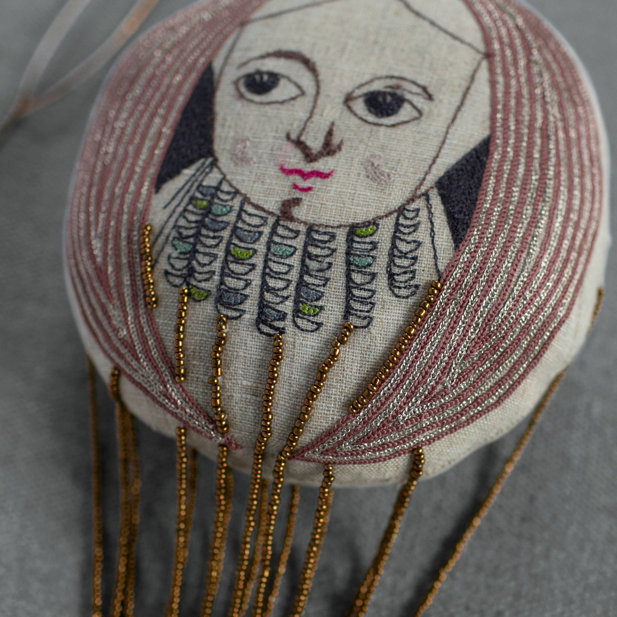 The Skippy Cotton "Wonderful Wizard / Balloon, Oz Wallhanging, Ornament, Token" is a round fabric piece with a stylized face, pink lips, long hair, gold bead strands, and decorative embroidery—a unique Wizard of Oz wall art item.