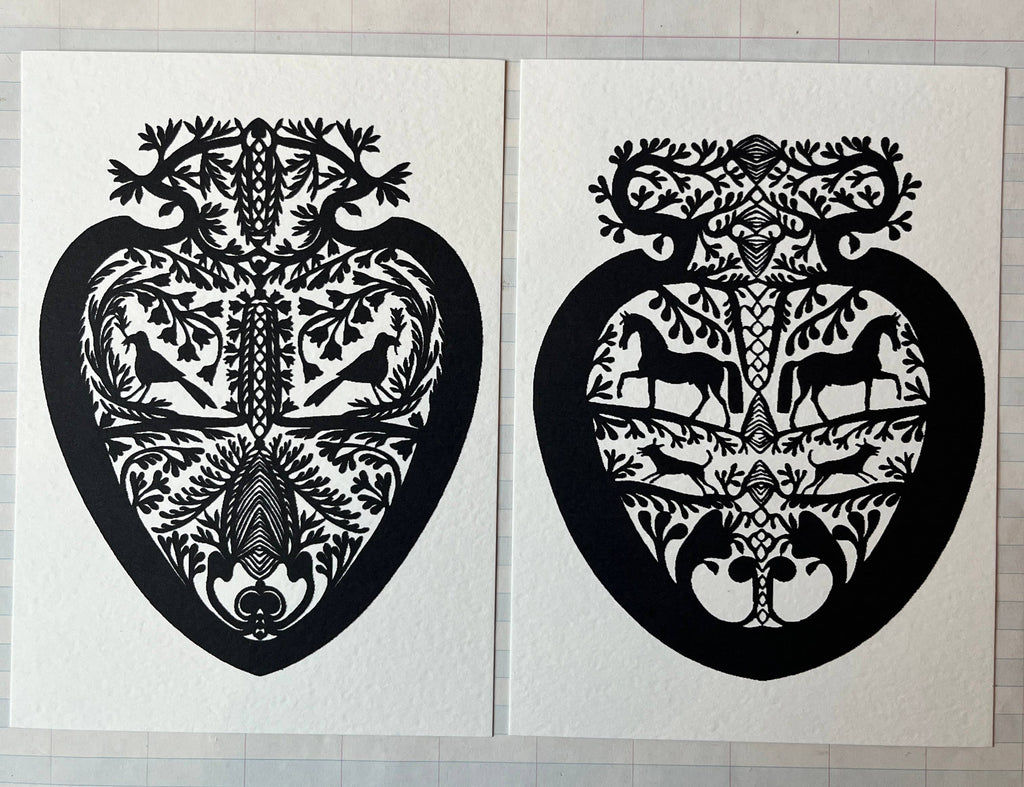 Parcel’s Papercut Heart Art Cards feature two 5x7 black heart-shaped designs with symmetrical birds, deer, and leaves. Displayed side by side on a grid-patterned background, each comes with a creamy white envelope.