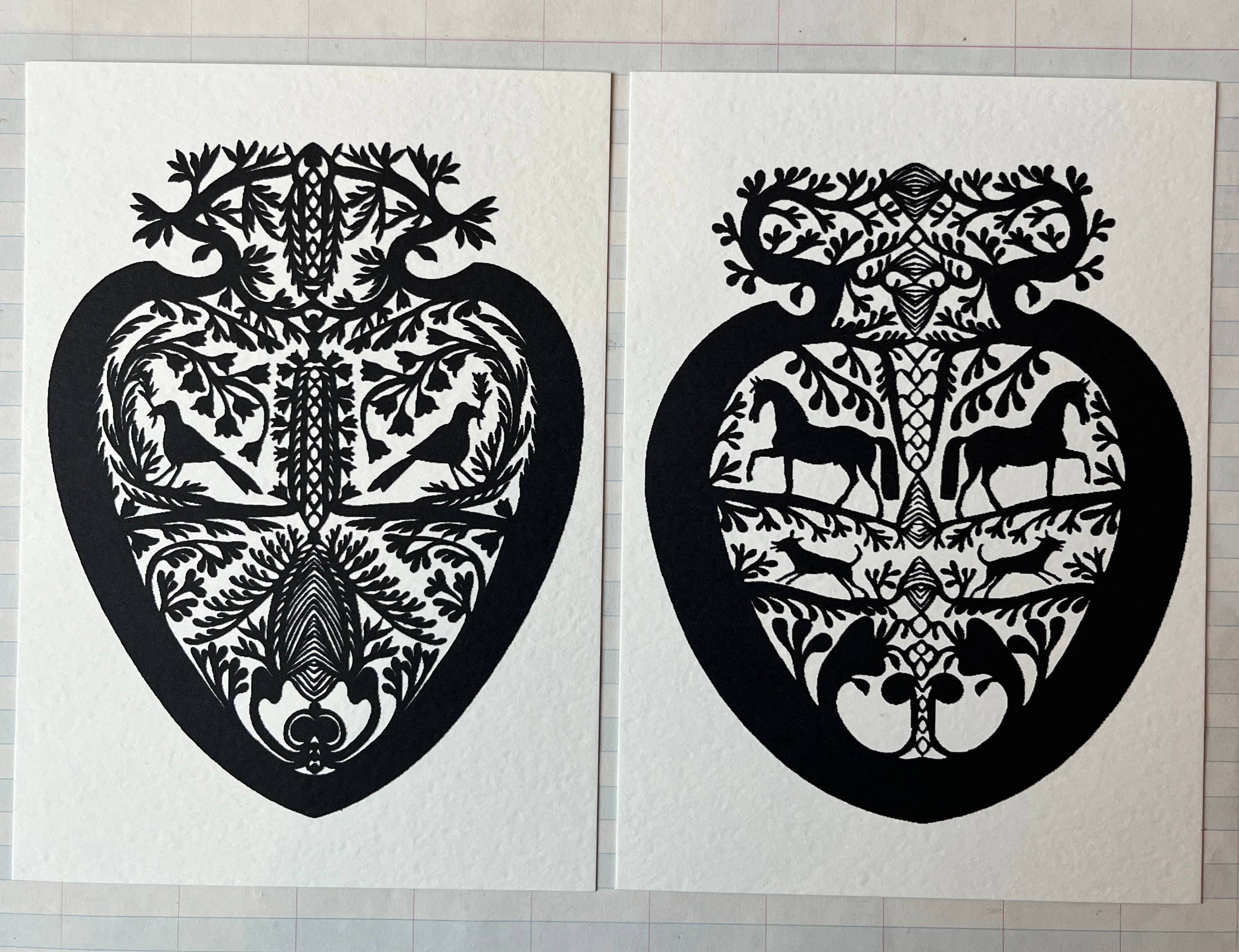 Parcel’s Papercut Heart Art Cards feature two 5x7 black heart-shaped designs with symmetrical birds, deer, and leaves. Displayed side by side on a grid-patterned background, each comes with a creamy white envelope.