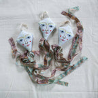 Three handmade Flying Kite ornaments by Skippy Cotton, crafted from organic cotton and filled with lavender. Each features illustrated faces with blue eyes, fabric ribbons, and is perfect as a scented gift topper or décor piece.
