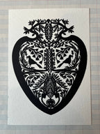 The Parcel Papercut Heart Art Card features a symmetrical black heart design with intricate birds, trees, and leaves, adorning a 5x7 card displayed on lined paper.