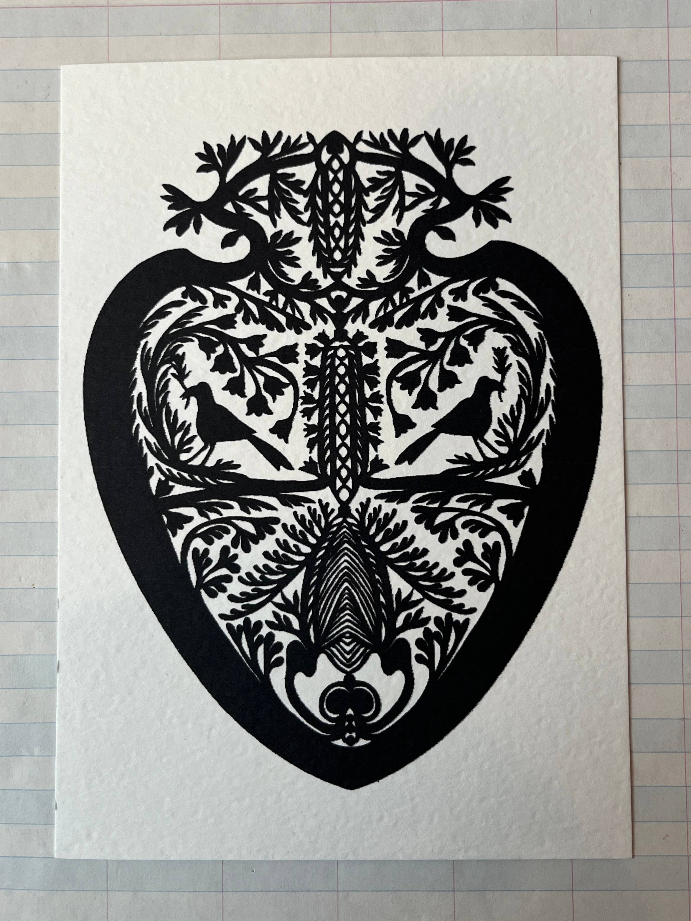 The Parcel Papercut Heart Art Card features a symmetrical black heart design with intricate birds, trees, and leaves, adorning a 5x7 card displayed on lined paper.