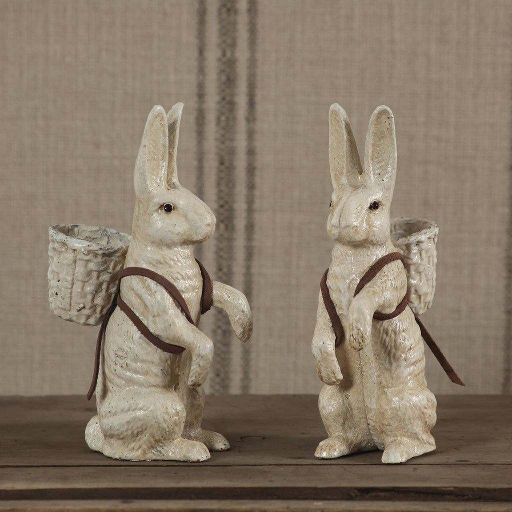 The HomArt Mr. Hamilton Cast Iron rabbit figurines in antique white stand upright on a wooden surface, each with a small woven basket and brown straps, set against a beige striped fabric—ideal for charming home decor.