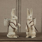 The HomArt Mr. Hamilton Cast Iron rabbit figurines in antique white stand upright on a wooden surface, each with a small woven basket and brown straps, set against a beige striped fabric—ideal for charming home decor.