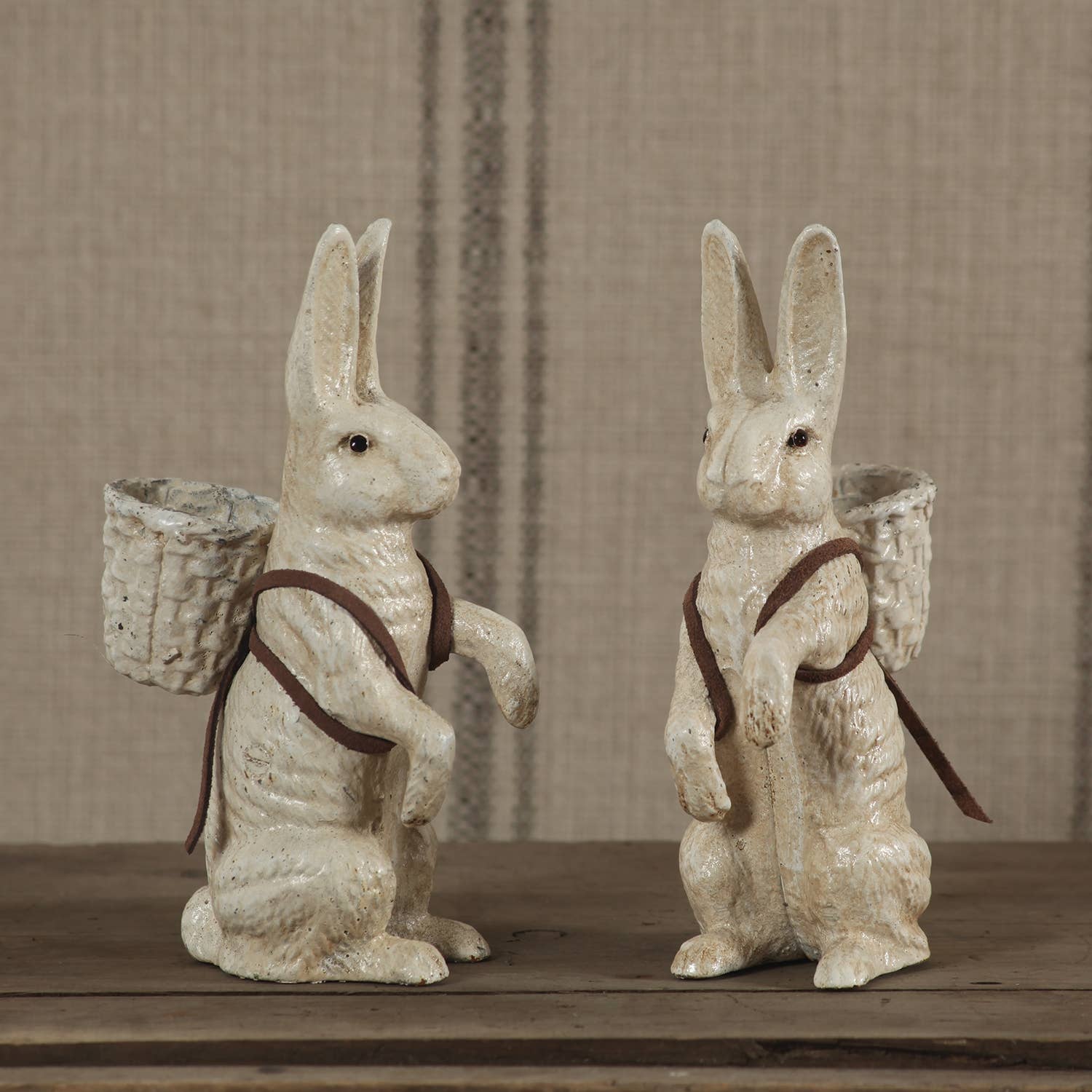 The HomArt Mr. Hamilton Cast Iron rabbit figurines in antique white stand upright on a wooden surface, each with a small woven basket and brown straps, set against a beige striped fabric—ideal for charming home decor.