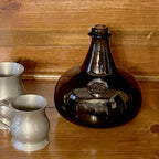A Hand-Blown Dark Green 76-oz. Onion Bottle, 8" by Madison Bay Co. sits on a wooden surface next to two pewter mugs, with a wooden wall in the background.