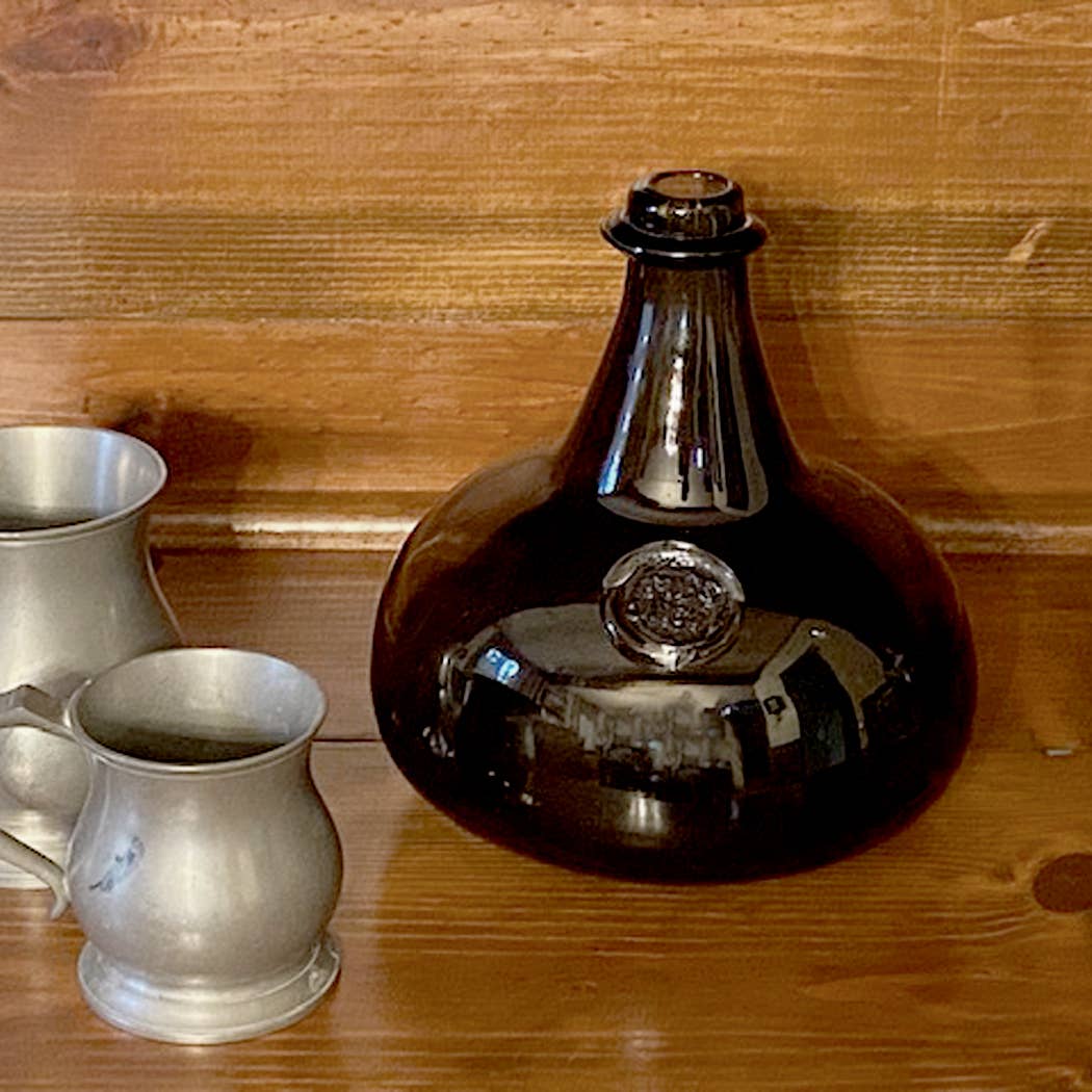 A Hand-Blown Dark Green 76-oz. Onion Bottle, 8" by Madison Bay Co. sits on a wooden surface next to two pewter mugs, with a wooden wall in the background.