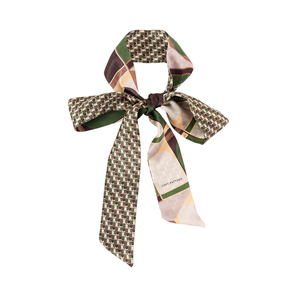 The Silk Skinny Scarf - Green by Lost Pattern NYC features a geometric pattern in green, brown, cream, and yellow. Made of silk satin with double-sided stitching and LOST PATTERN branding—ideal as a chic hair accessory tied in a bow.